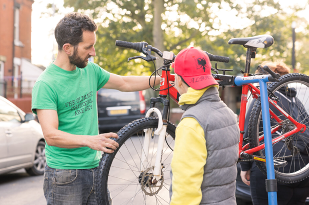 A community repair café volunteer demonstrates bike maintenance to a visitor outdoors. The session, supported by enfinium’s Repair Café Support Fund, highlights sustainable living practices and waste reduction by teaching skills to extend the life of everyday items like bicycles.