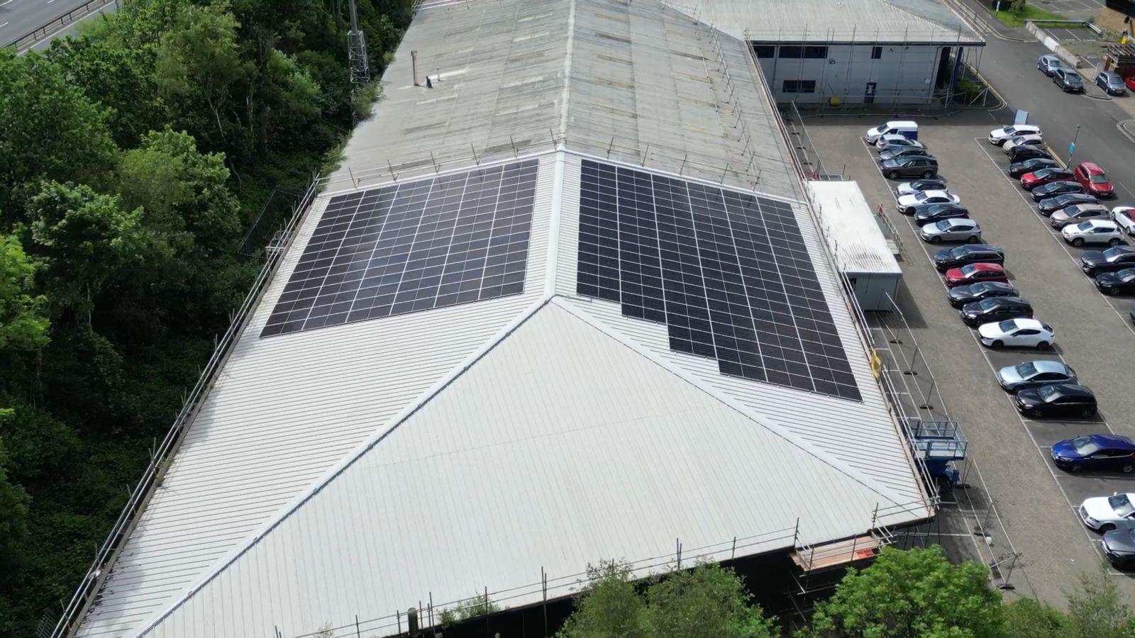 Aerial view of a warehouse with a large solar panel installation on its roof, designed to maximise energy efficiency and sustainability. The panels cover a significant portion of the roof, and the surrounding area features a parking lot with several vehicles. This installation demonstrates the potential of solar energy to enhance warehouse operations and reduce carbon footprints.