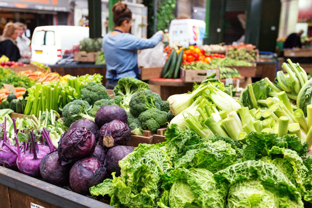 An abundance of fresh, locally sourced vegetables at a market stall, showcasing the potential of sustainable nutrition through seasonal and low-impact produce, with a vendor actively engaged in supporting a circular food economy.