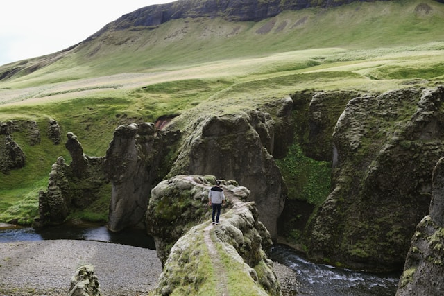 Person standing on a narrow grassy cliff surrounded by dramatic moss-covered rock formations and a winding river in a remote Icelandic valley.