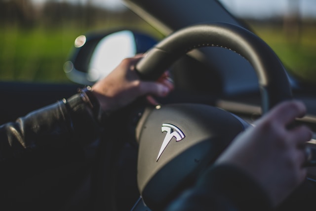 Close-up of a person holding the steering wheel of a Tesla while driving, with a blurred background visible through the window.