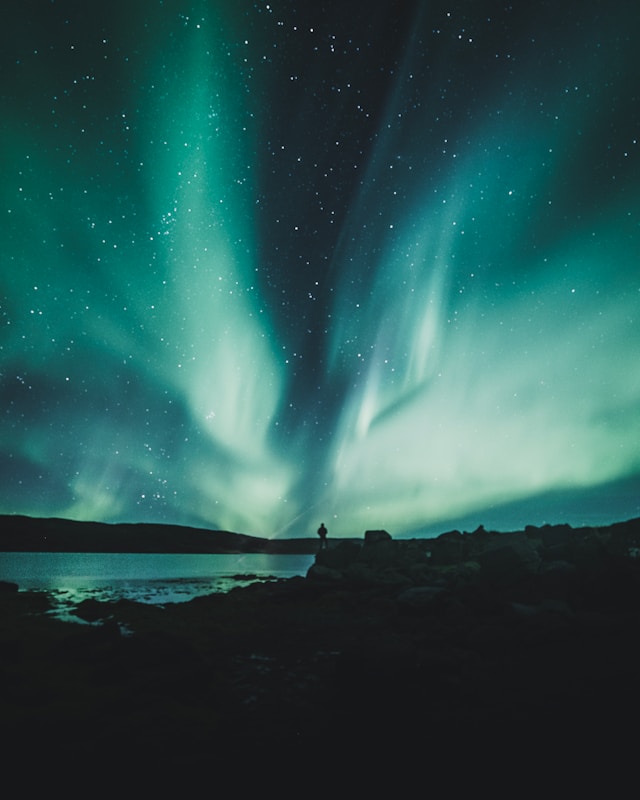 Person standing near a serene body of water under a vibrant display of the Northern Lights, representing the natural beauty and sustainability focus of Iceland ecotourism.