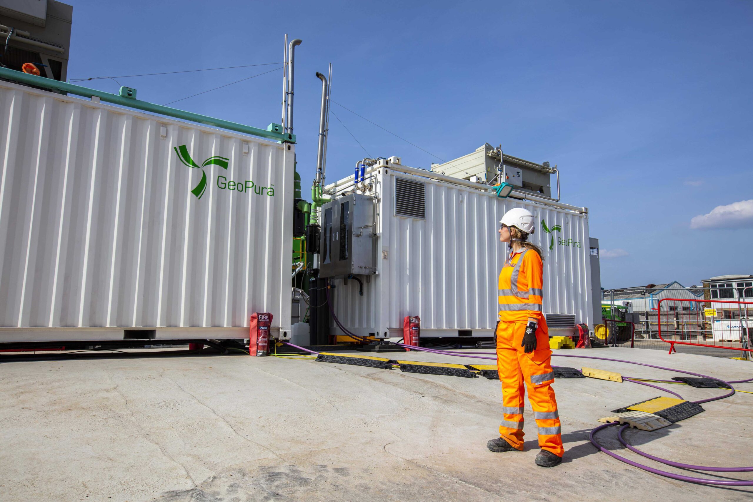 A worker in high-visibility orange protective gear and a white hard hat stands in front of a GeoPura hydrogen generator unit at an industrial site, with cables and safety equipment visible on the ground under a clear blue sky.