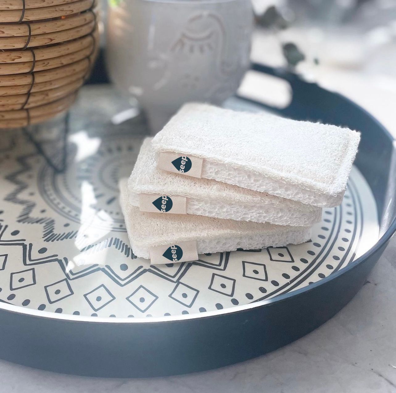 Three eco-friendly, reusable white sponges with small brand tags stacked neatly on a decorative round tray with a geometric pattern. The background features a woven basket, a ceramic teapot, and a softly blurred setting, giving a clean and minimalist aesthetic.