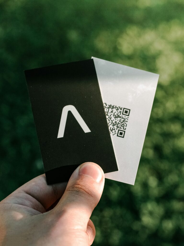 Hand holding two modern business cards eco friendly, one with a minimalist logo and the other featuring a QR code, against a natural green background.