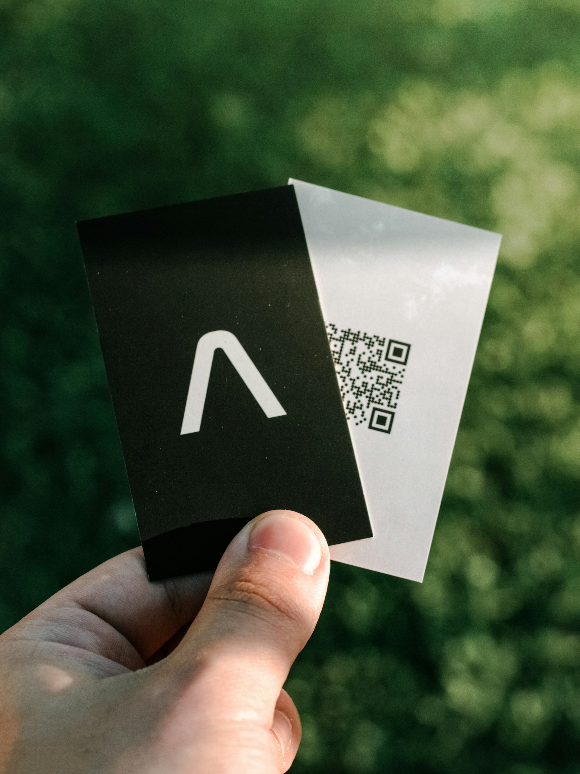 Hand holding two modern business cards eco friendly, one with a minimalist logo and the other featuring a QR code, against a natural green background.