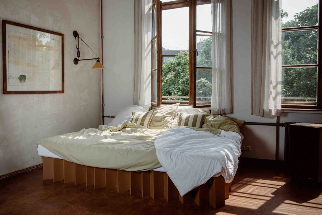 A minimalist bedroom featuring a sustainable cardboard bed frame with a white and light green bedding set. The room has large open windows with sheer curtains, allowing natural light to fill the space. The walls are textured with a rustic finish, and a framed artwork hangs on the left side. A vintage-style wall lamp extends over the bed, complementing the eco-friendly and modern aesthetic.