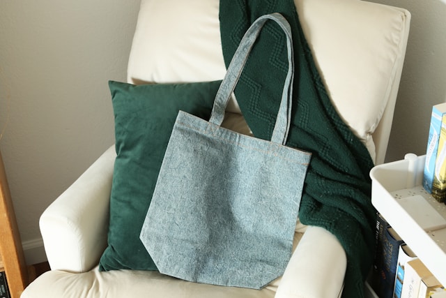 A light blue denim tote bag resting on a white armchair with a green pillow and throw blanket - an excellent example of what to do with old jeans by transforming them into functional, stylish accessories.