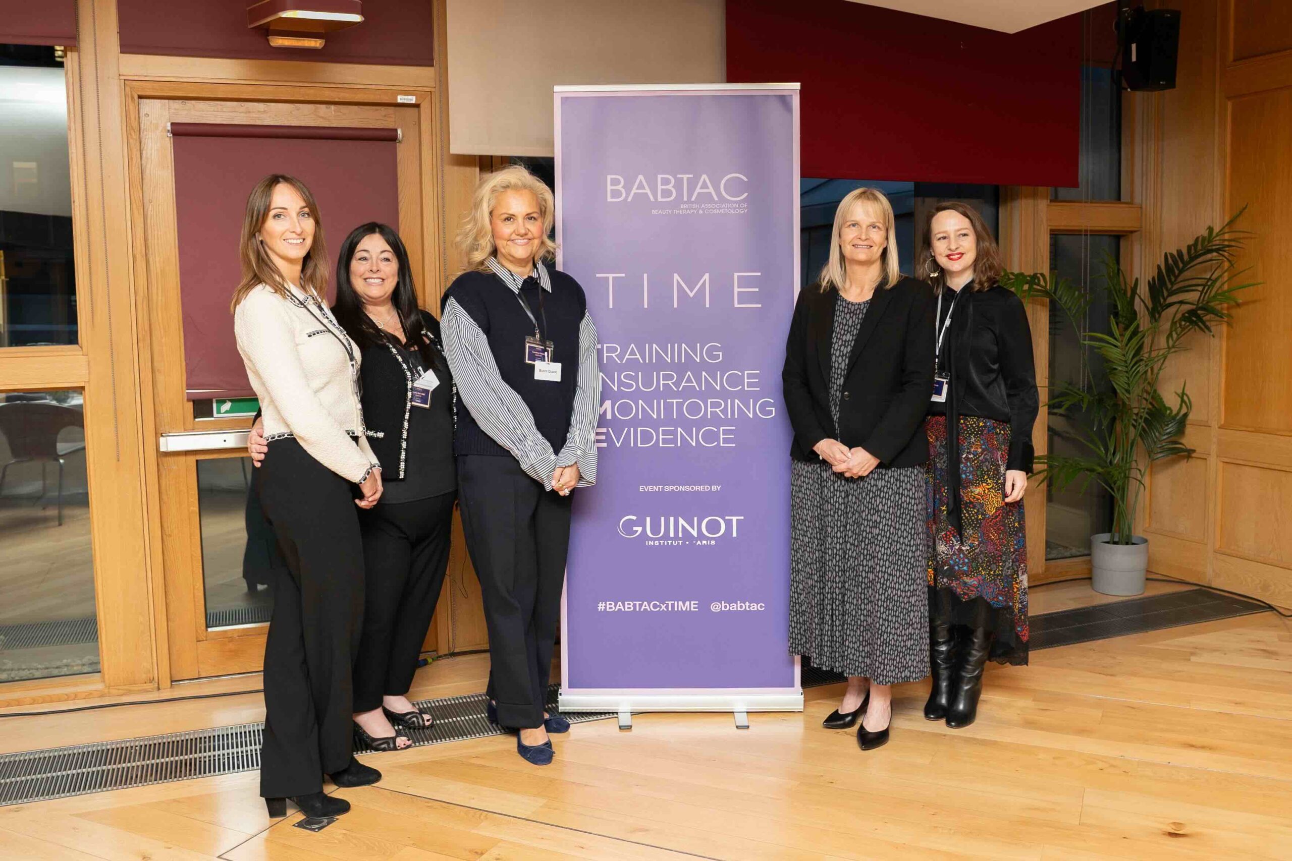 A group of five women standing in front of a Beauty Industry Regulation in Scotland banner at a BABTAC T.I.M.E initiative event. The banner highlights key principles: Training, Insurance, Monitoring, and Evidence. The event, sponsored by Guinot, takes place in an indoor venue with wooden paneling and potted plants.