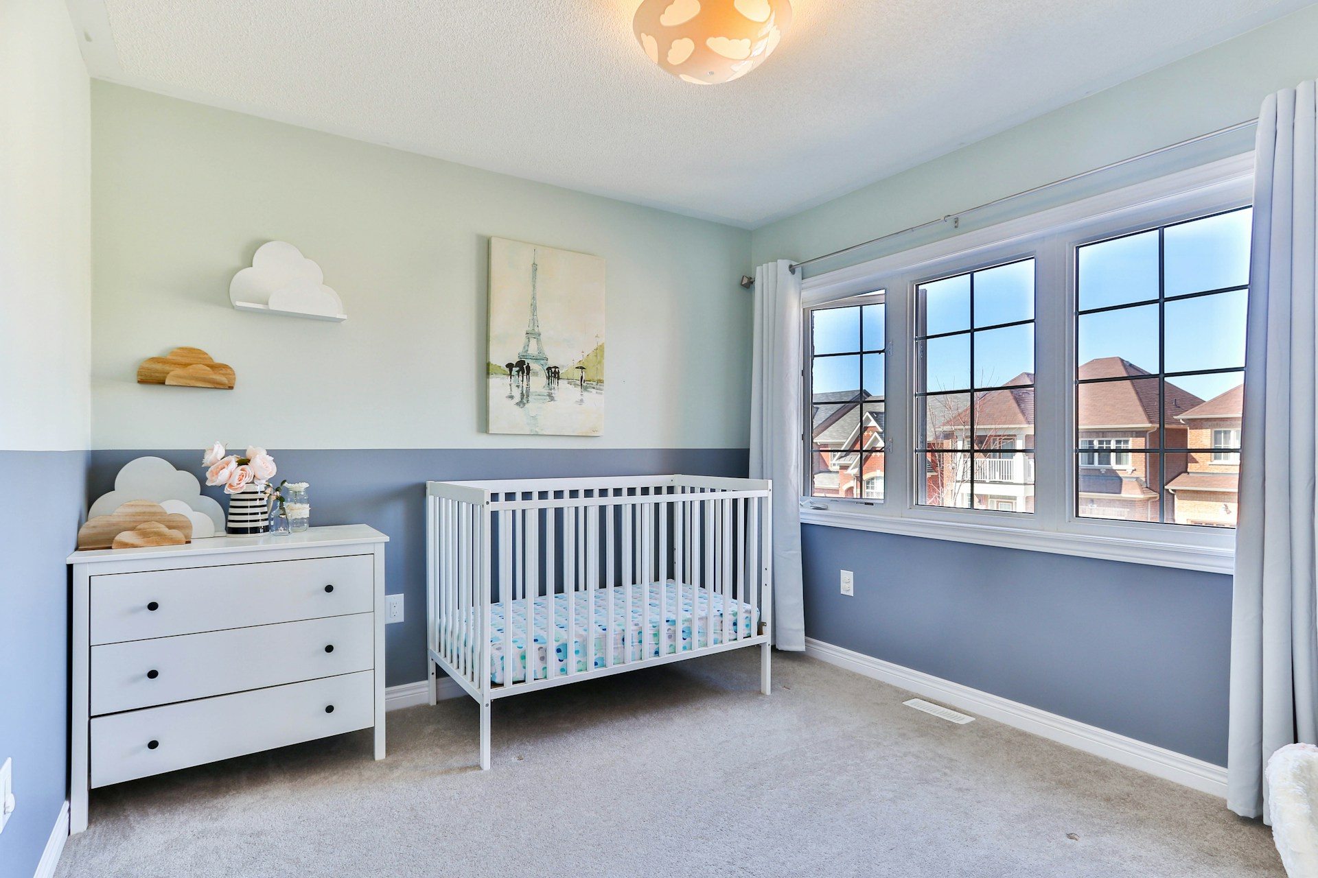 A modern nursery featuring a white crib painted with the best non-toxic paint, alongside a matching white dresser, soft blue and gray walls, and cloud-themed décor.