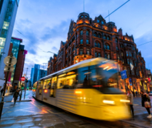 A motion-blurred yellow tram moving through Manchester city centre at dusk, with a historic red-brick building and modern skyscrapers in the background. Pedestrians walk along the wet streets under a vibrant evening sky.