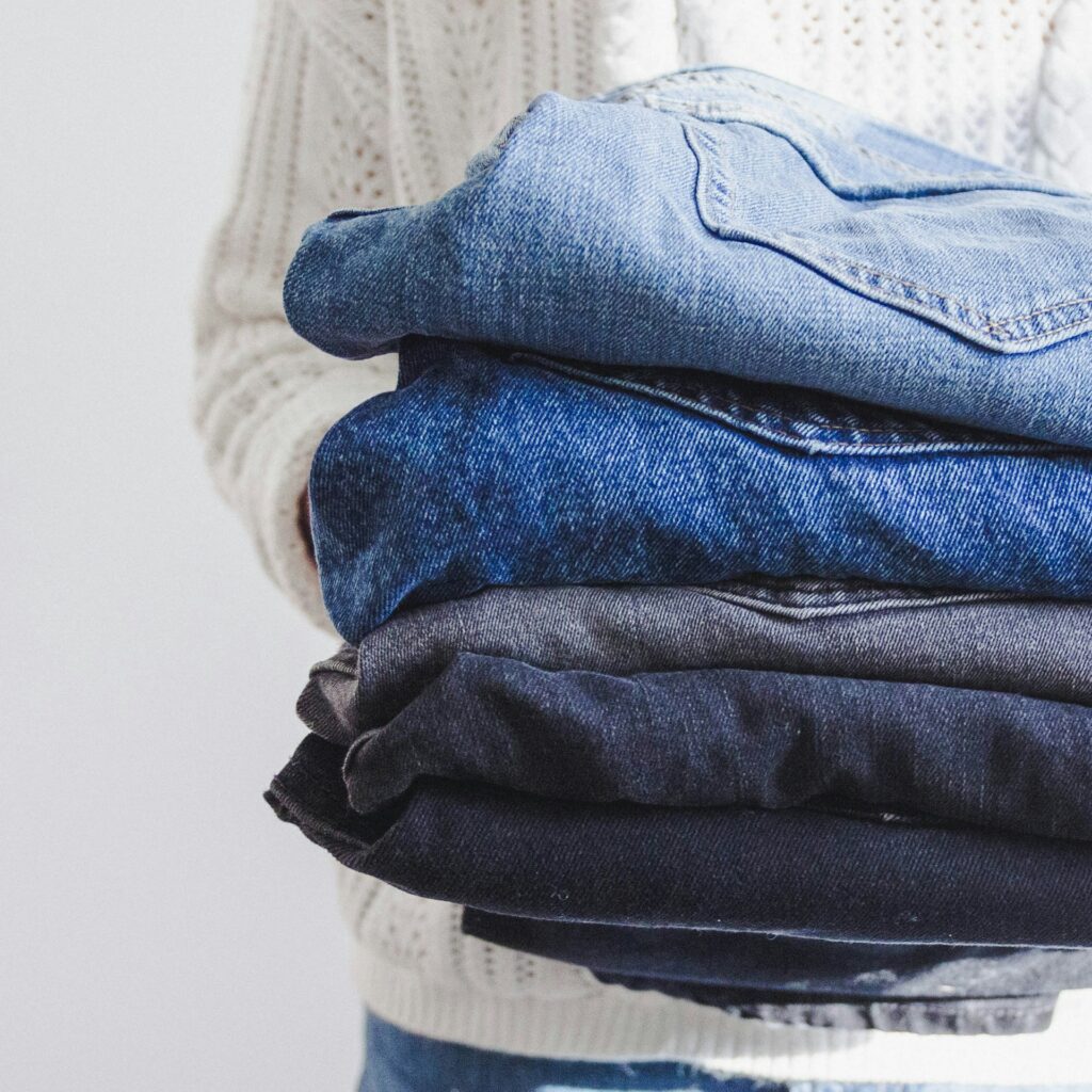 A person in a cream-colored sweater holding a neatly folded stack of denim jeans in varying shades of blue and black - perfect inspiration for what to do with old jeans when looking for sustainable fashion projects.