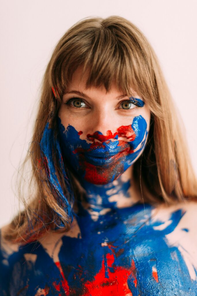 Woman with red and blue body paint non toxic applied across her face and upper body, smiling confidently in front of a light background.