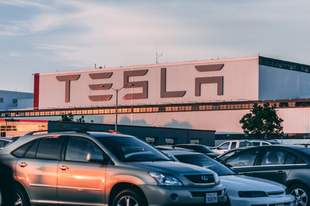 Tesla factory building with cars parked in front, capturing the scale of electric vehicle production and sparking the question: why can't electric cars charge themselves?