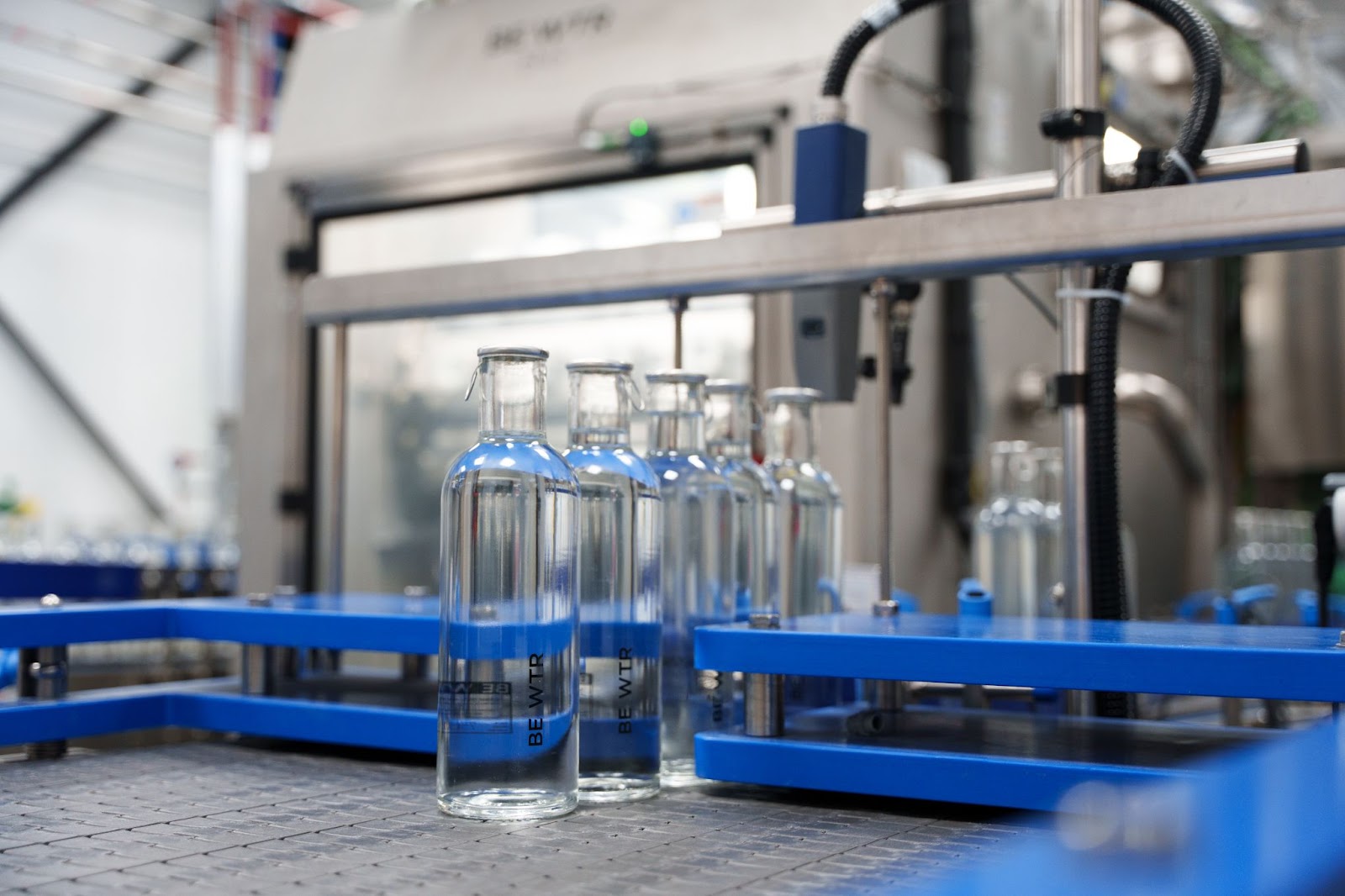 Reusable glass water bottles labeled "BE WTR" being filled on an automated production line at a sustainable bottling plant. The process highlights eco-friendly water solutions in a modern industrial setting.