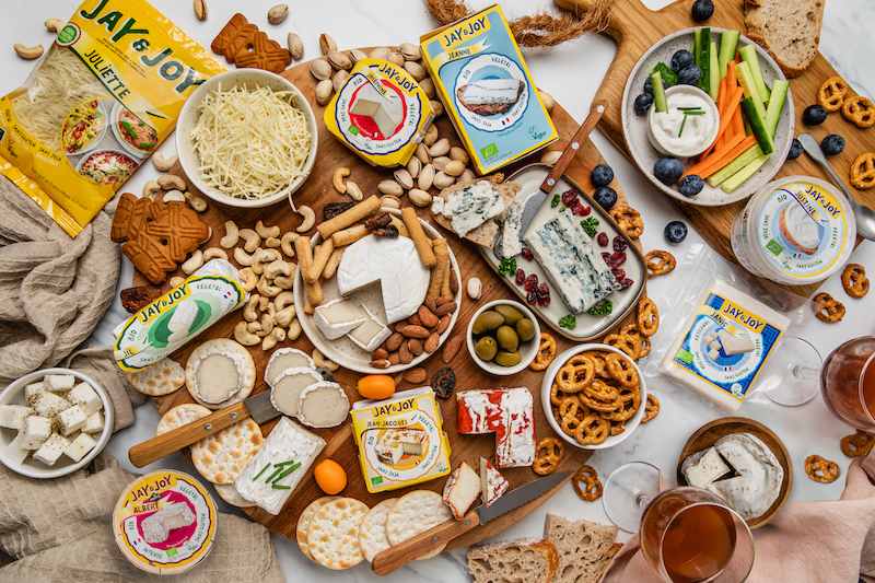 A vibrant vegan cheeseboard featuring a variety of Jay&Joy plant-based cheeses, fresh vegetables, crackers, olives, nuts, and fruits arranged on a wooden board.