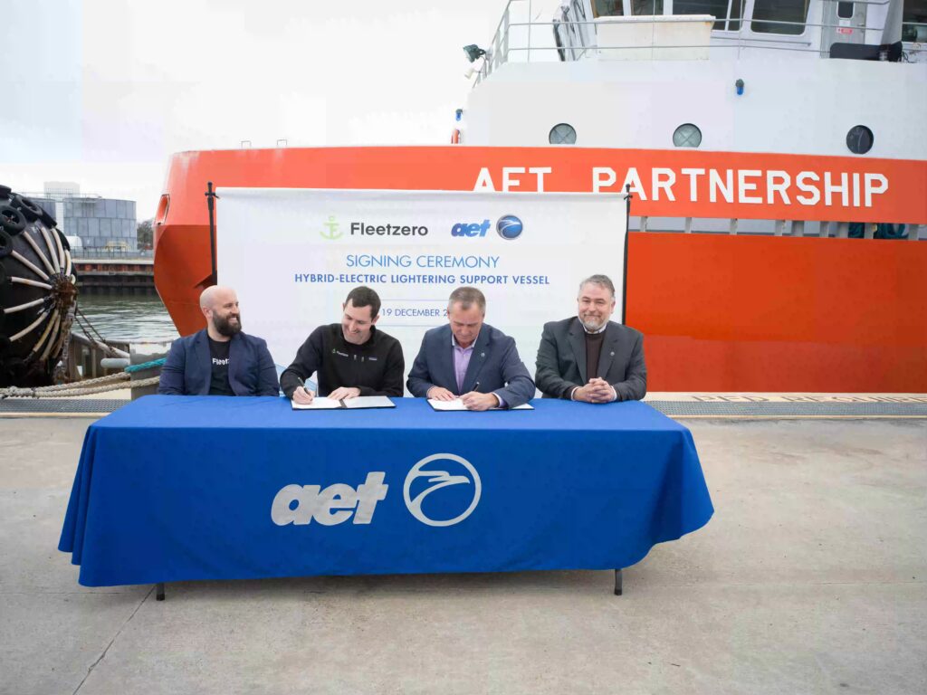 Representatives from AET and Fleetzero signing an agreement for the Longest-Range Hybrid Electric Vessel, seated at a table in front of an AET partnership backdrop.