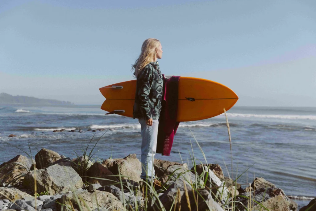 Woman holding a surfboard on a rocky coastline, wearing outdoor sustainable clothing by the ocean.