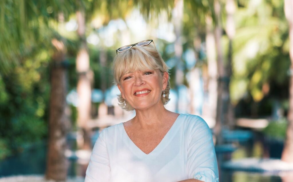 Narelle McDougall, General Manager of Amilla Maldives, a sustainable luxury resort in the Maldives, smiling outdoors with tropical palm trees in the background.
