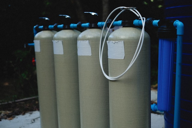 A row of large industrial water filtration tanks connected by blue PVC pipes, used in a residential or commercial water treatment setup.







