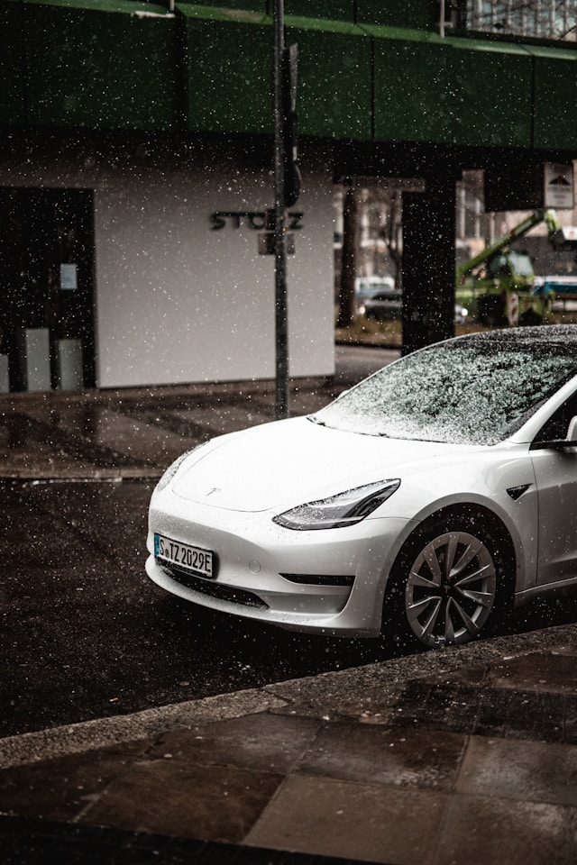 White Tesla Model 3 parked on a rainy city street, with water droplets covering the car and surroundings.