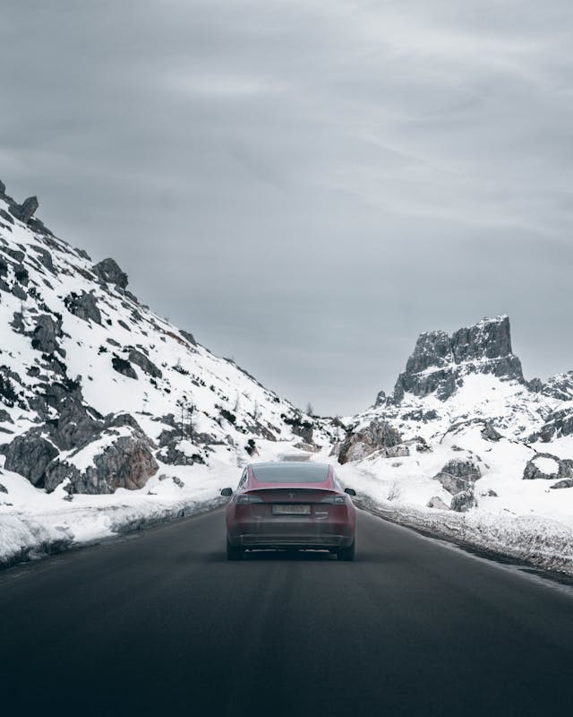 Red Tesla driving on a mountain road surrounded by snow and rocky peaks under a cloudy sky.
