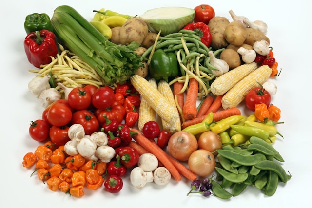 A colorful assortment of fresh produce including carrots, corn, tomatoes, mushrooms, peppers, and leafy greens arranged on a white background.