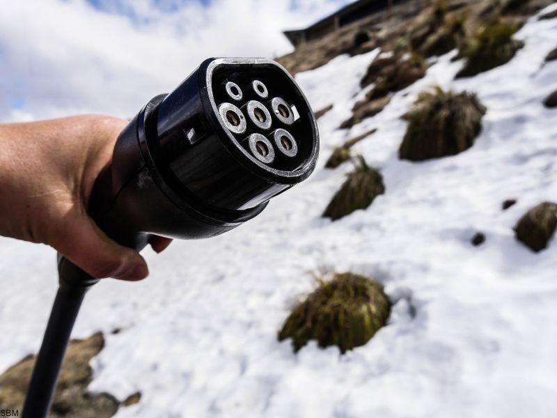 A close-up of a person holding an electric vehicle charging plug in a snowy mountain environment, symbolizing the question of how much electricity does a Tesla use in different conditions
