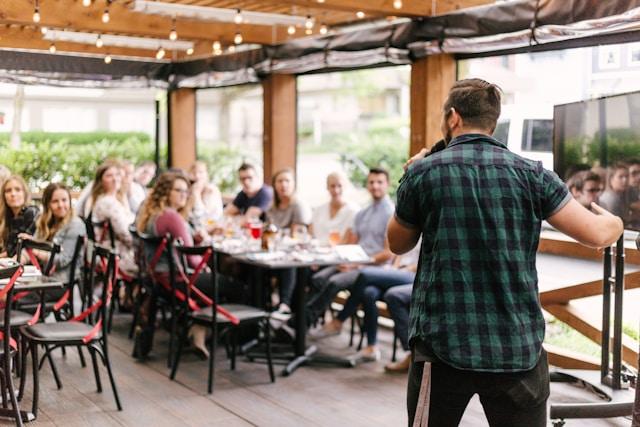 A speaker addresses a diverse group of young professionals seated at an outdoor restaurant-style gathering, symbolizing community engagement and training opportunities often associated with entry level jobs in nonprofit organizations, where communication and collaboration are essential.