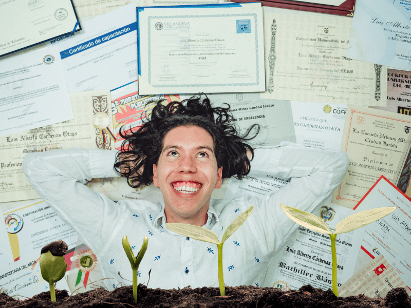 Smiling man lying on a floor covered with certificates, with seedlings growing in soil in the foreground, symbolizing achievement and growth through sustainability certifications.