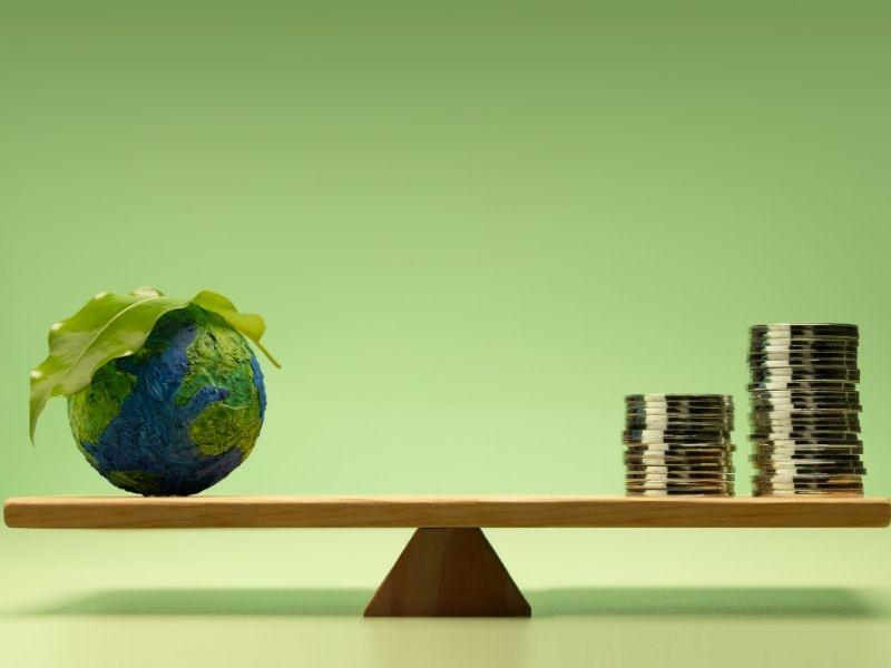 A conceptual image symbolizing ecological business, showing a miniature Earth with a green leaf on top balanced on one side of a wooden seesaw, while stacks of coins rest on the other side. The background is a solid light green, emphasizing sustainability and the balance between environmental responsibility and economic growth.