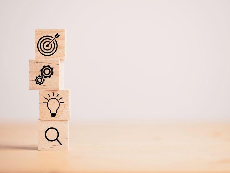 Stacked wooden blocks with icons of a target, gears, lightbulb, and magnifying glass, symbolizing goal-setting and planning in ethical investing.