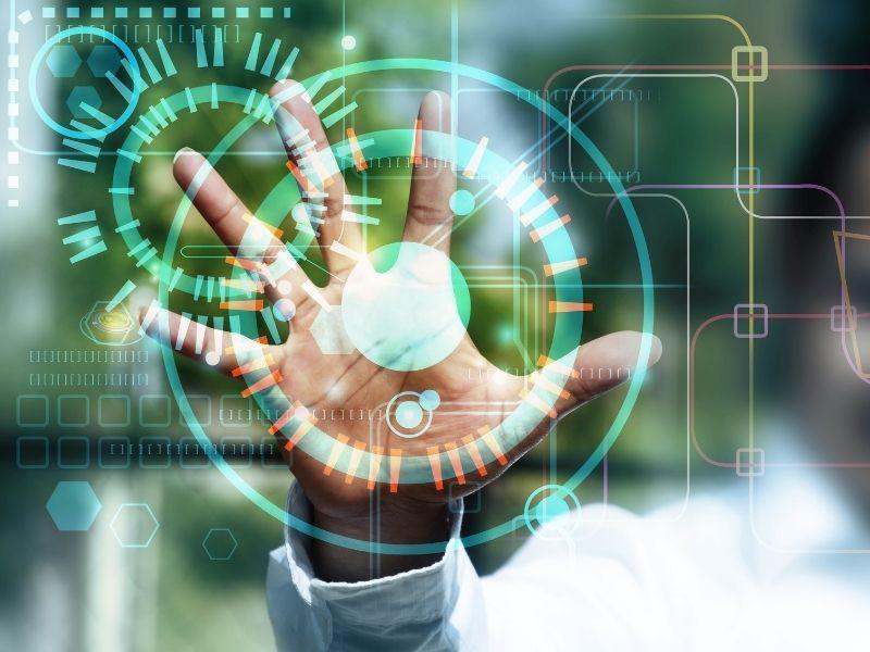A human hand reaching out toward a transparent, futuristic interface filled with circular graphs, digital indicators, and glowing data elements. The background is a blurred natural scene, suggesting a blend of technology and nature. The image visually represents the integration of eco-friendly technologies for sustainable living through advanced digital solutions.