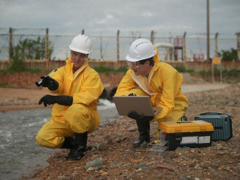 Two environmental scientists wearing yellow protective suits and white hard hats collect water samples and analyze pollution data near an industrial site, illustrating the challenges and complexity of studying environmental science and fieldwork.