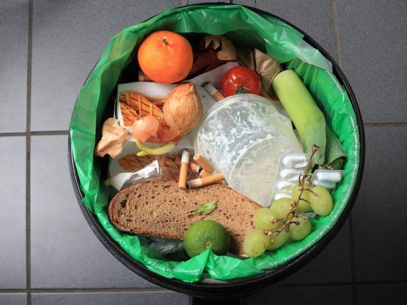Household food waste and plastic trash in a green bin, showing bread, fruits, vegetables, and packaging, highlighting food waste and recycling issues.