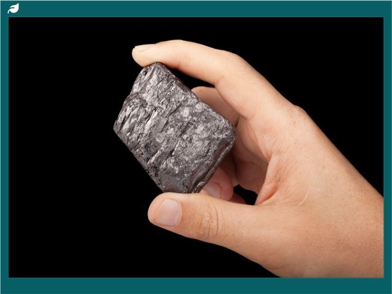 Hand holding a raw piece of carbon, highlighting carbon properties such as hardness, metallic luster, and natural texture against a black background.
