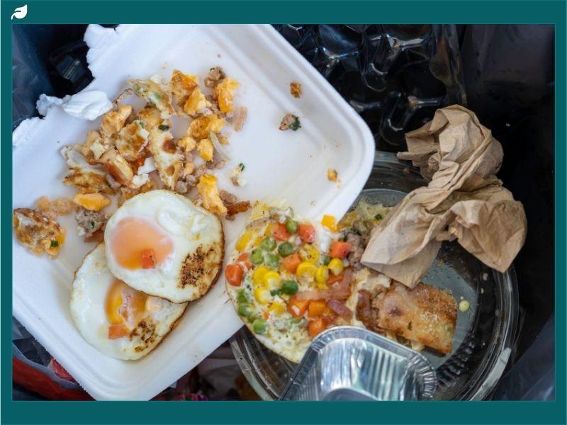 A discarded takeout container with uneaten fried eggs, mixed vegetables, and bread thrown into the trash, emphasizing the importance of actions to reduce food waste.