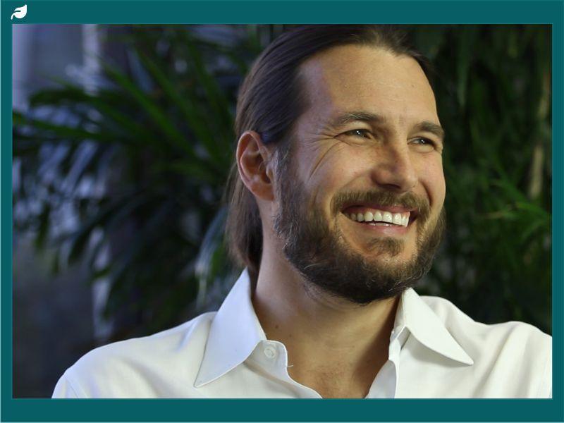 A smiling man with long hair and a beard wearing a white shirt, seated in front of green foliage, conveying warmth and openness.