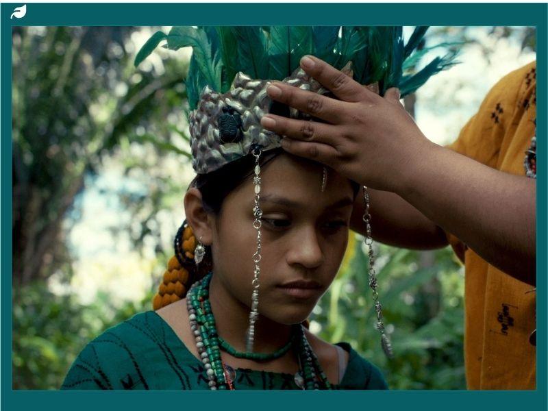 A young person wearing traditional clothing and jewelry being crowned with a feathered headdress during a cultural ceremony, set outdoors among lush greenery.
