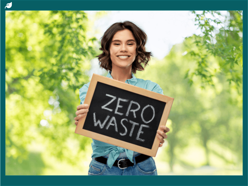 A smiling woman outdoors holding a chalkboard sign that says “Zero Waste,” promoting the zero waste lifestyle and sustainable living practices.