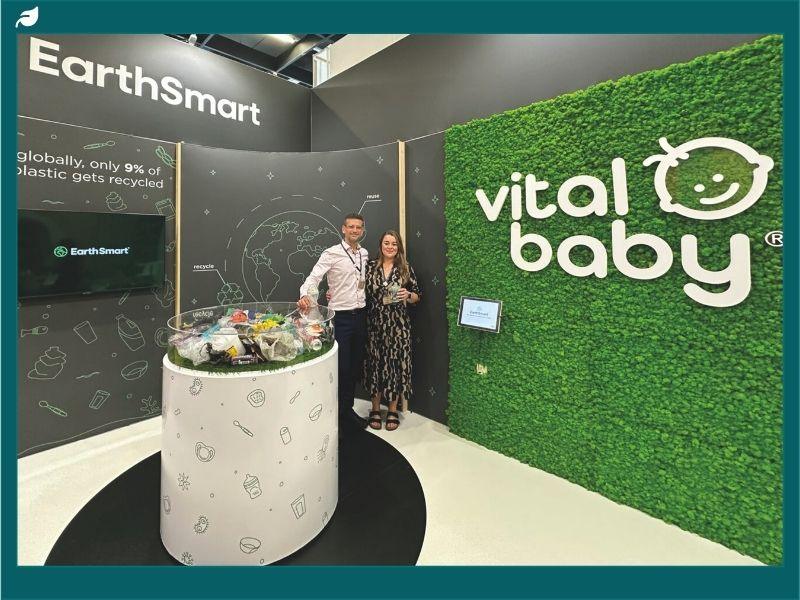 Two people stand at a Vital Baby EarthSmart exhibition booth with green logo wall and a display table filled with plastic products