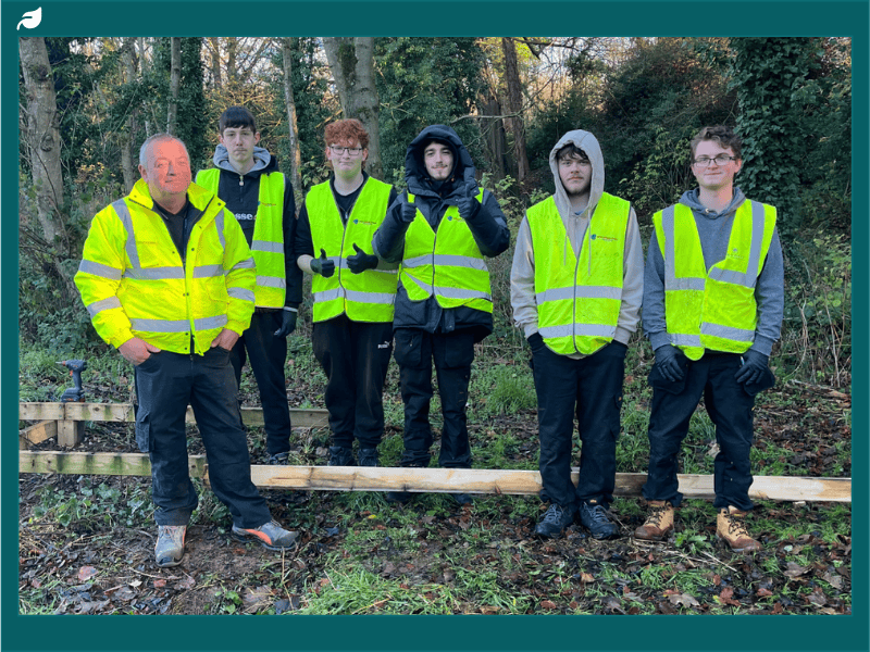 Stephenson College carpentry students and instructor wearing high-visibility vests standing in woodland at Snibston Colliery Park during the boardwalk regeneration project.