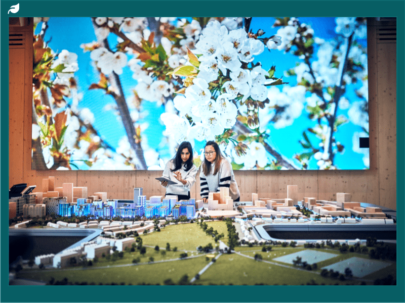 Two ladies at Ramboll working on a model of a town. They stand in front of a large picture of white flowers. 