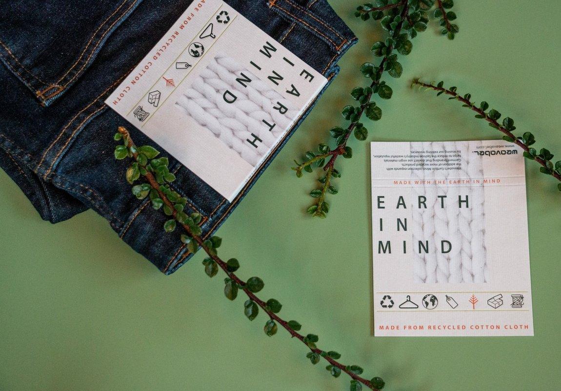 Dark blue jeans with sustainable "EARTH IN MIND" tag and matching card displayed with green foliage on light green background