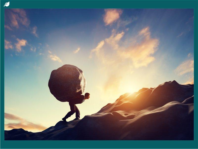 Silhouetted man struggling to carry massive boulder up steep hill