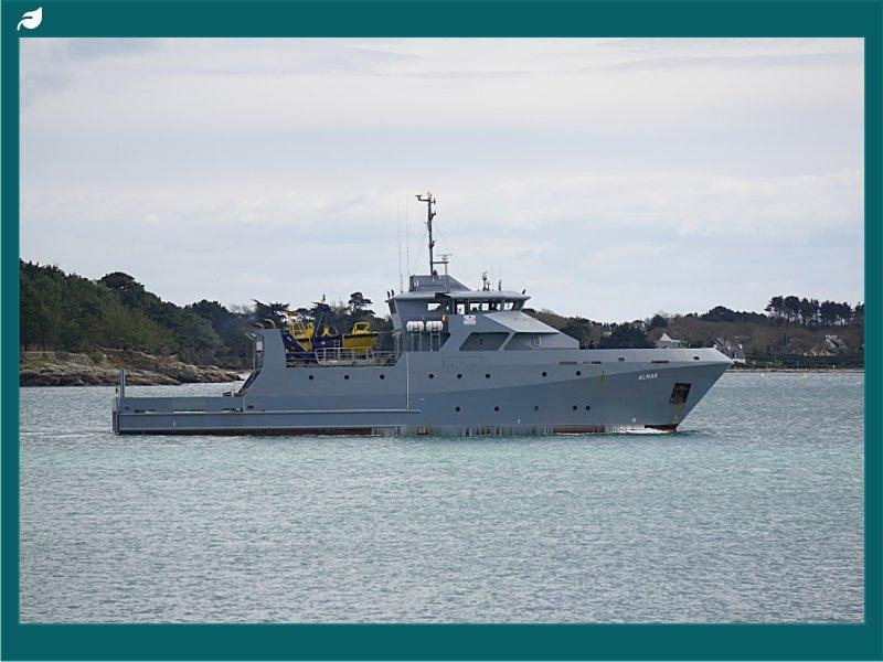 Grey offshore support vessel named Alnair sailing along a wooded coastline