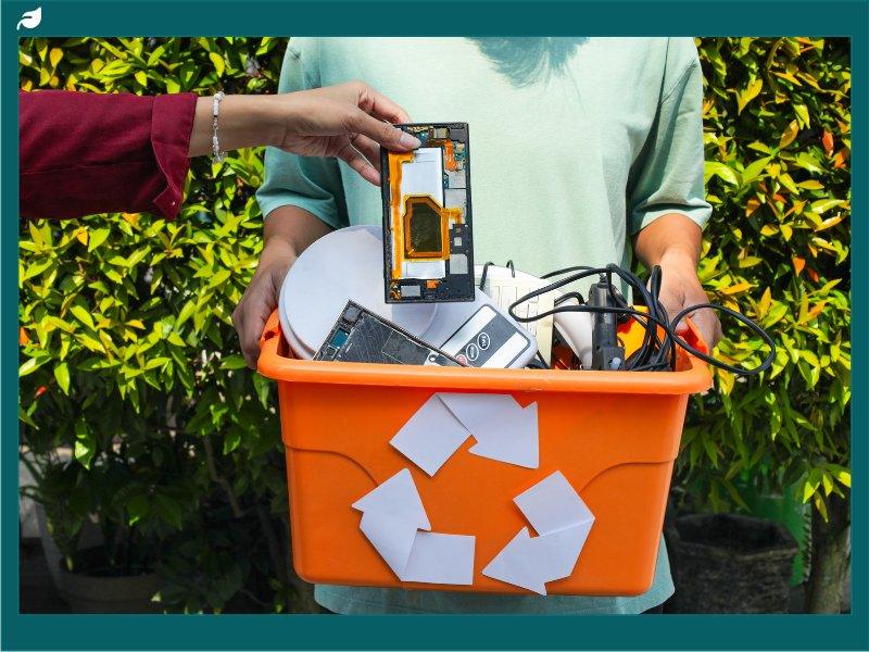 Person Places Broken Smartphone Component Into Recycling Bin Filled With Assorted Electronic Waste Held By Another Individual Outdoors