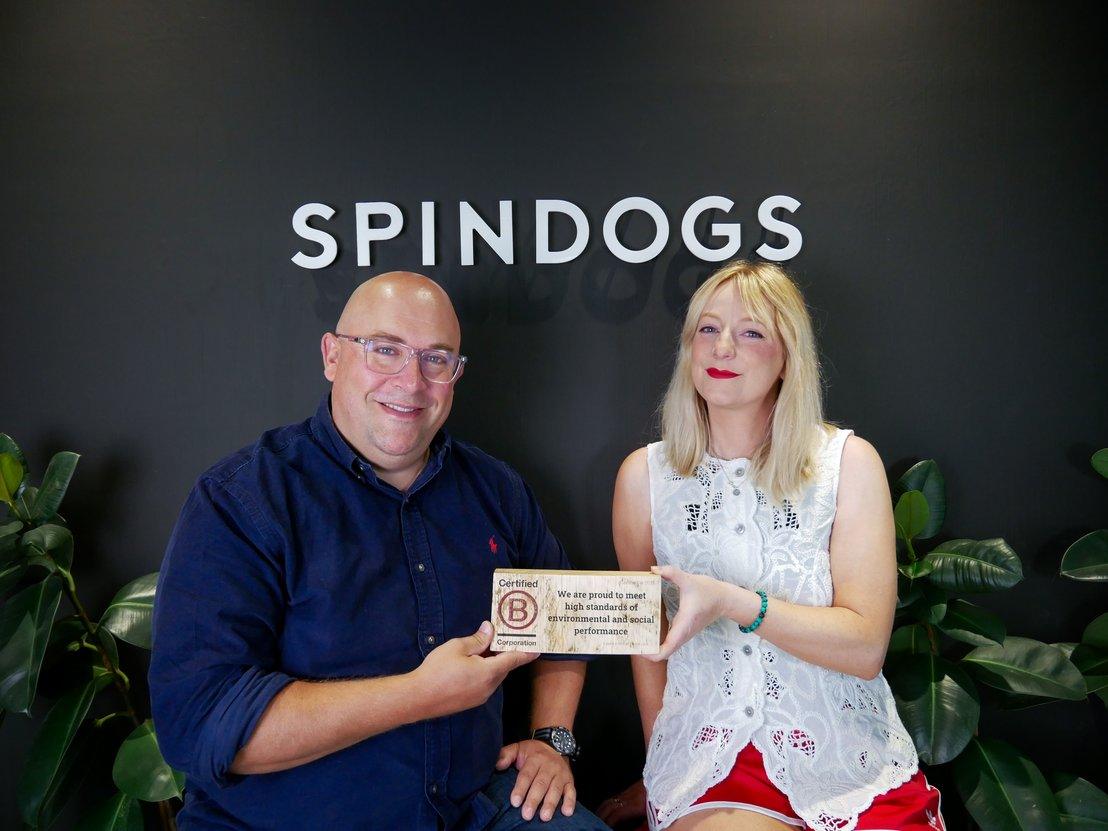 Two Spindogs employees holding a B Corp certification plaque in front of the Spindogs logo wall