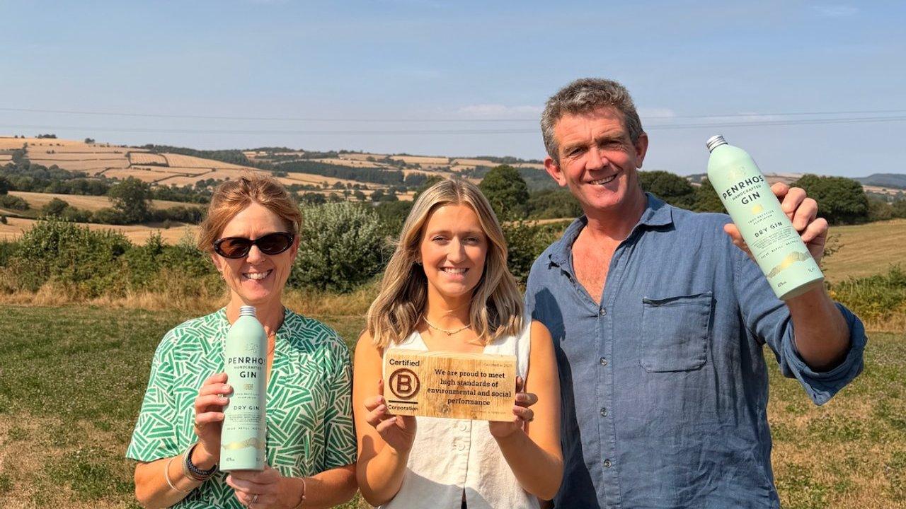 Three people hold gin bottles and a B Corporation plaque in a rural landscape.