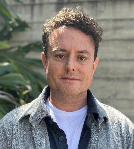 A man with curly brown hair wearing a grey jacket outdoors beside leafy plants and a concrete wall